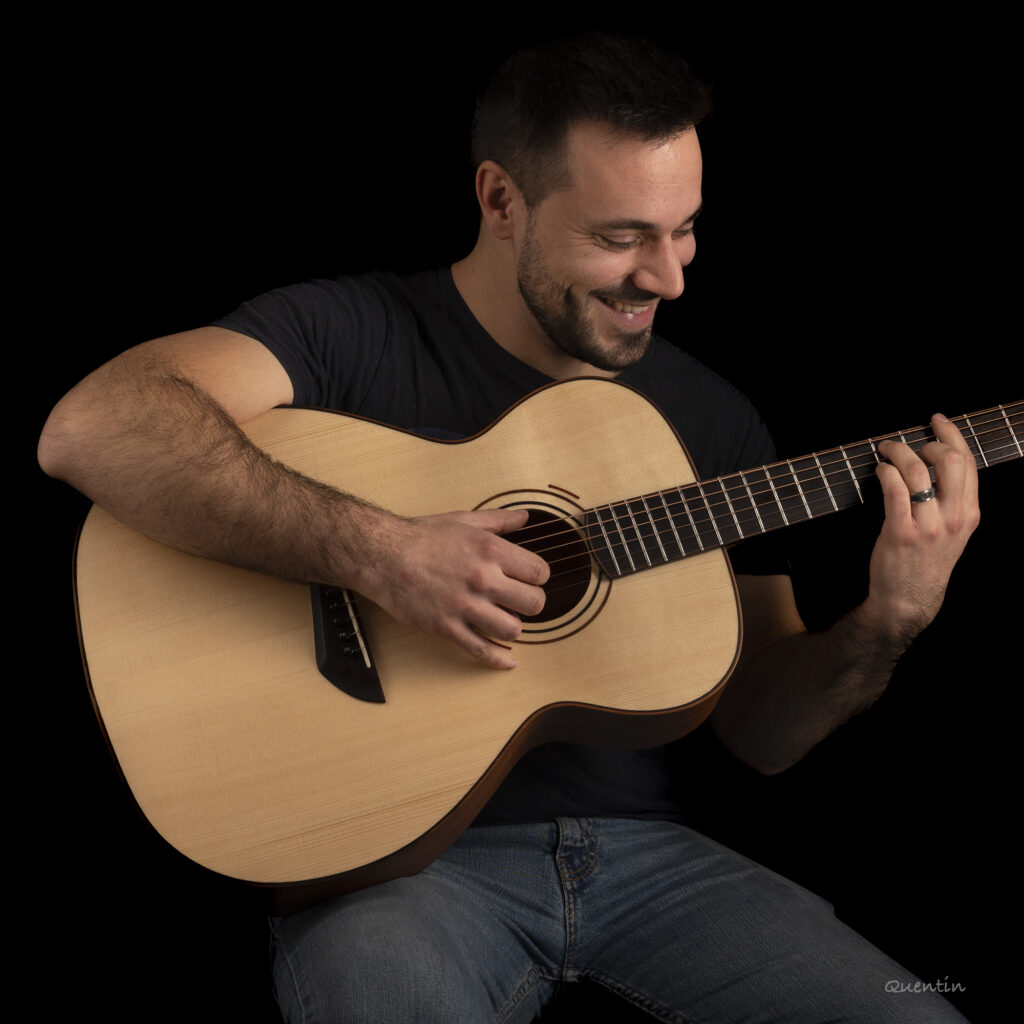 Le luthier sur fond noir avec une de ses guitares, il en joue et se montre souriant.